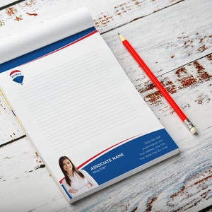 May include: A notepad with the RE/MAX logo, a red and blue design, and lined pages. A red pencil with a white eraser rests beside the notepad. The notepad features a photo of a woman and the words "Associate Name, Realtor."