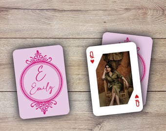 May include: A pink card with a decorative pink frame and the text "E Emily". A white playing card with a picture of a woman sitting on a chair and the text "Queen of Hearts" is behind the pink card.