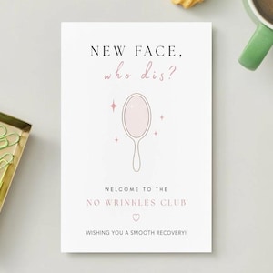 Könnte beinhalten: Eine Grußkarte mit dem Text "NEW FACE, who dis?" und "WELCOME TO THE NO WRINKLES CLUB". Die Karte zeigt eine Handspiegel-Illustration und ist von einem goldenen Tablett mit grünen Büroklammern und einer Tasse Kaffee umgeben.