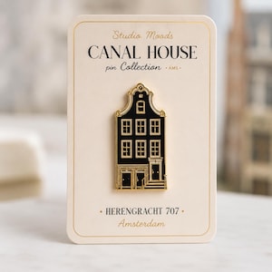 Op de afbeelding: Een zwart-gouden emaille speld in de vorm van een grachtenpand, bevestigd op een crèmekleurige kaart. De kaart bevat de tekst Studio Moods CANAL HOUSE pin Collection AMS en HERENGRACHT 707 Amsterdam in zwarte schreef- en scriptlettertypes.