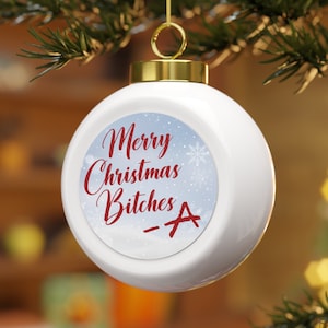 May include: White Christmas ornament with a gold cap and hanger. The ornament features the text "Merry Christmas Bitches -A" in red script on a blue and white background with snowflakes. The ornament is hanging from a green branch.
