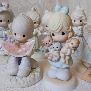 May include: A collection of Precious Moments figurines. The figurines depict children in pastel-colored outfits, holding items like teddy bears, watermelon, and flowers. The figurines have large, expressive eyes and are set on decorative bases.