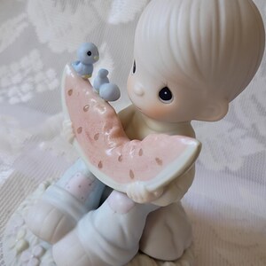 May include: A porcelain figurine of a child holding a watermelon slice, with a small blue bird perched on top. The figurine has a soft, pastel color palette and is set on a decorative base. The child's clothing is light blue and pink.
