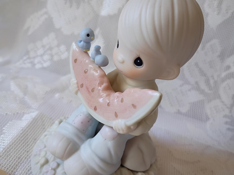 May include: A porcelain figurine of a child holding a watermelon slice, with a small blue bird perched on top. The figurine has a soft, pastel color palette and is set on a decorative base. The child's clothing is light blue and pink.