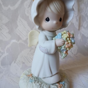 May include: A porcelain figurine of an angel, wearing a white dress and a halo. The angel holds a bouquet of pastel-colored flowers. The figurine stands on a base with painted grass and small flowers.