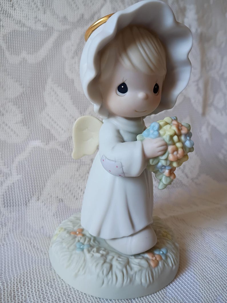 May include: A porcelain figurine of an angel, wearing a white dress and a halo. The angel holds a bouquet of pastel-colored flowers. The figurine stands on a base with painted grass and small flowers.