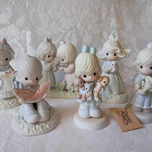 May include: A collection of Precious Moments figurines. The figurines depict children in pastel colors, some holding animals or objects. The figurines are on a white lace tablecloth. A tag with the Precious Moments logo is visible.