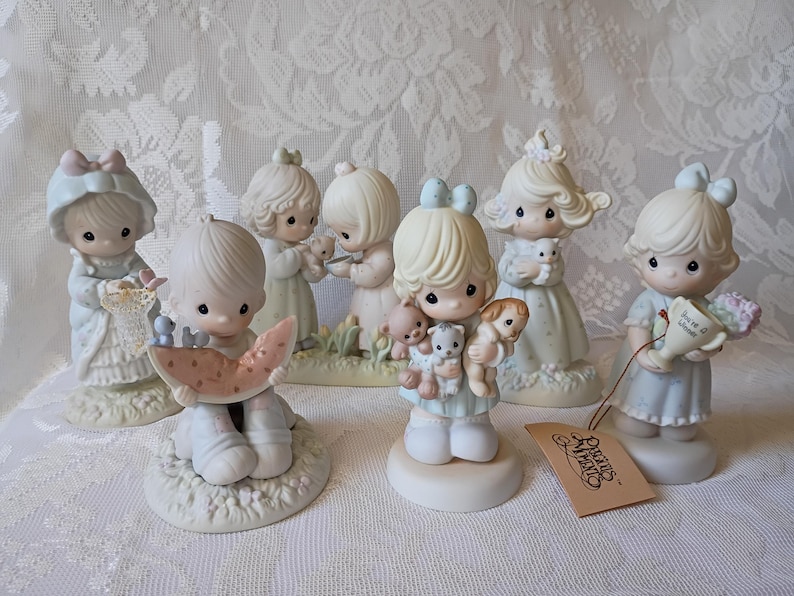 May include: A collection of Precious Moments figurines. The figurines depict children in pastel colors, some holding animals or objects. The figurines are on a white lace tablecloth. A tag with the Precious Moments logo is visible.