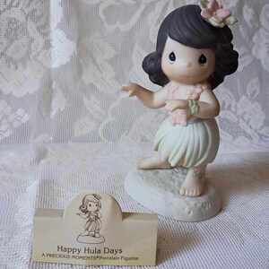 May include: A porcelain figurine of a girl in a hula skirt, with a flower in her hair and a lei. The figurine is on a small base. A small card reads "Happy Hula Days".