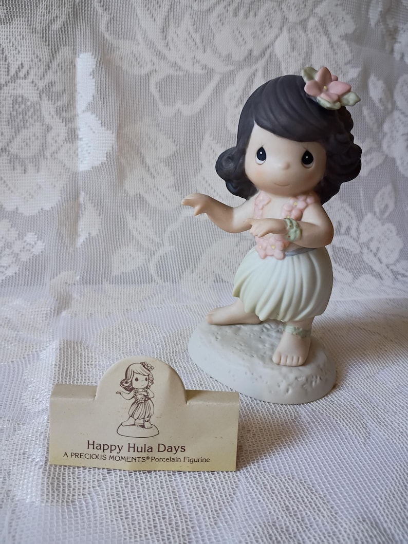 May include: A porcelain figurine of a girl in a hula skirt, with a flower in her hair and a lei. The figurine is on a small base. A small card reads "Happy Hula Days".
