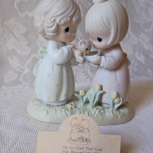 May include: A Precious Moments porcelain figurine featuring two figures in pastel dresses, holding a small bear and a bowl. The base is light green with small flowers. A card in front reads "I'm So Glad That God Has Blessed Me With A Friend Like You."