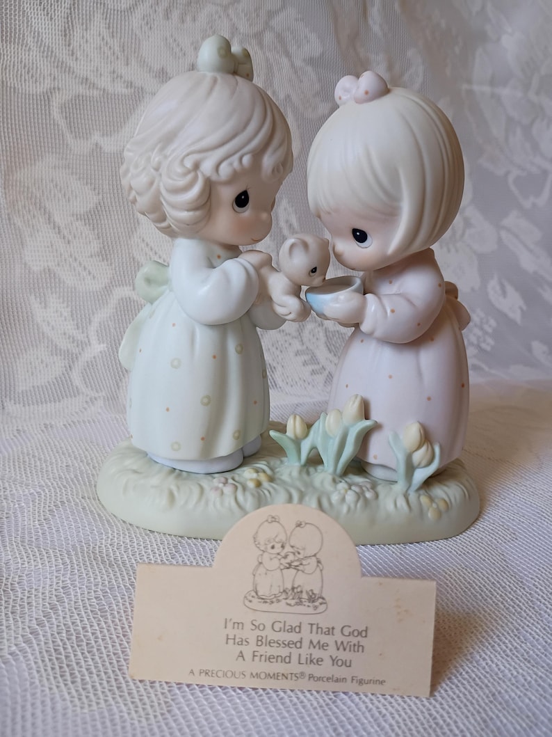 May include: A Precious Moments porcelain figurine featuring two figures in pastel dresses, holding a small bear and a bowl. The base is light green with small flowers. A card in front reads "I'm So Glad That God Has Blessed Me With A Friend Like You."