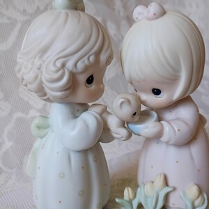 May include: A porcelain figurine set featuring two figures in pastel dresses, one holding a small bear and the other a blue bowl. The figures have blonde hair and are looking at the bear and bowl. The base has small flower details.