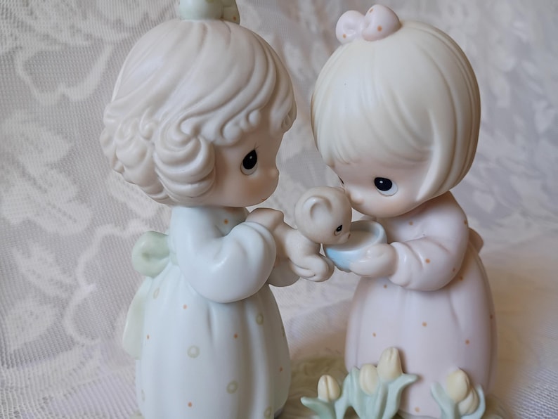 May include: A porcelain figurine set featuring two figures in pastel dresses, one holding a small bear and the other a blue bowl. The figures have blonde hair and are looking at the bear and bowl. The base has small flower details.