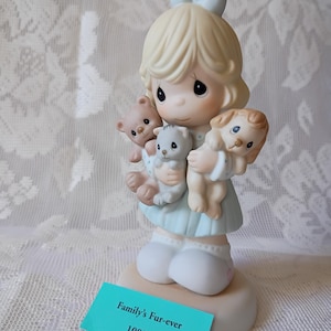 May include: A figurine of a young girl holding a teddy bear, a kitten, and a puppy. The girl wears a light blue dress and has a light blue bow in her hair. The figurine is on a round base with a teal tag that reads "Family's Fur-ever" and "108526".