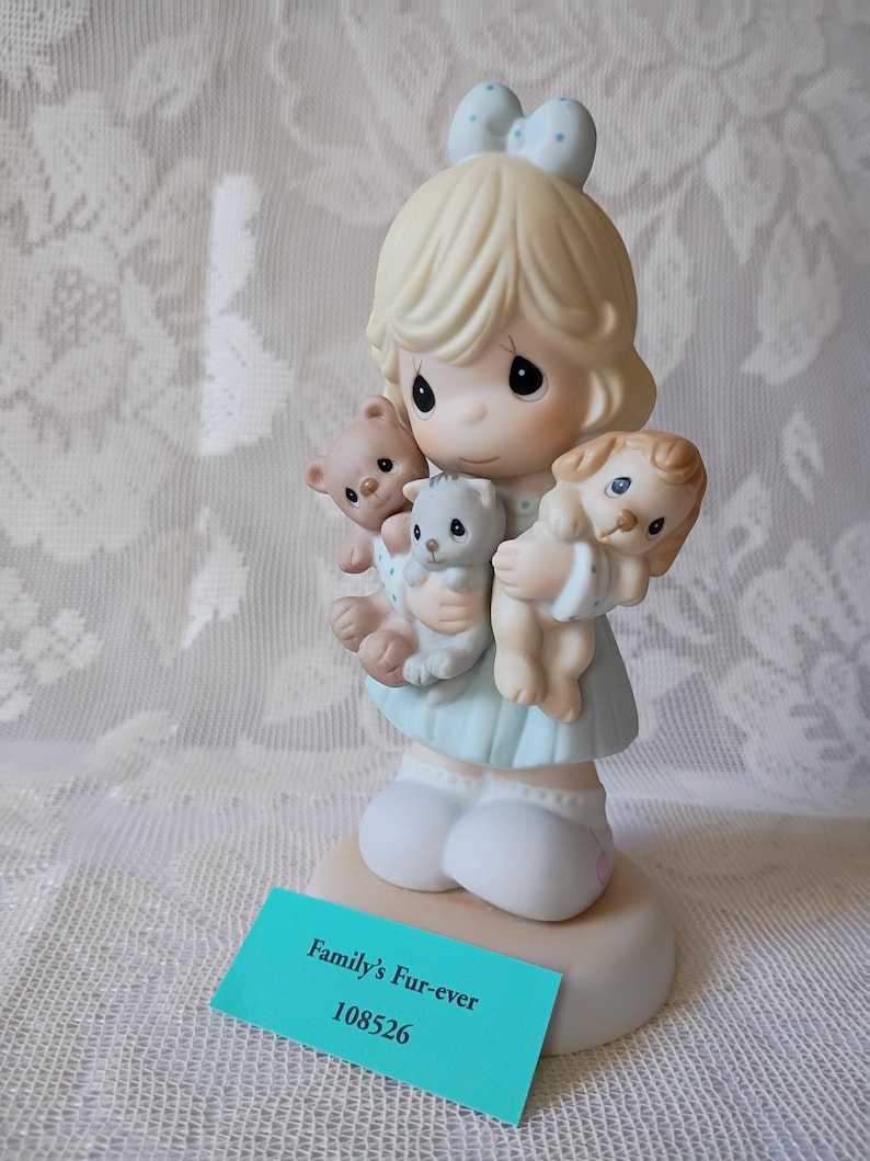 May include: A figurine of a young girl holding a teddy bear, a kitten, and a puppy. The girl wears a light blue dress and has a light blue bow in her hair. The figurine is on a round base with a teal tag that reads "Family's Fur-ever" and "108526".
