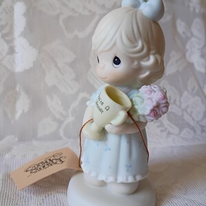 May include: A Precious Moments figurine of a girl in a light blue dress holding a trophy that says "You're A Winner" and a bouquet of pink flowers. The figurine has a light blue bow in her hair and is standing on a white base.