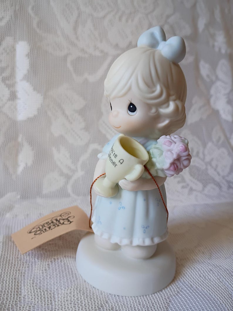 May include: A Precious Moments figurine of a girl in a light blue dress holding a trophy that says "You're A Winner" and a bouquet of pink flowers. The figurine has a light blue bow in her hair and is standing on a white base.