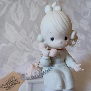 May include: A porcelain figurine of a girl with a bow in her hair, holding a pink telephone receiver. She is wearing a light blue dress with small polka dots. A small pink telephone sits on a white table. The figurine is part of the Precious Moments collection.