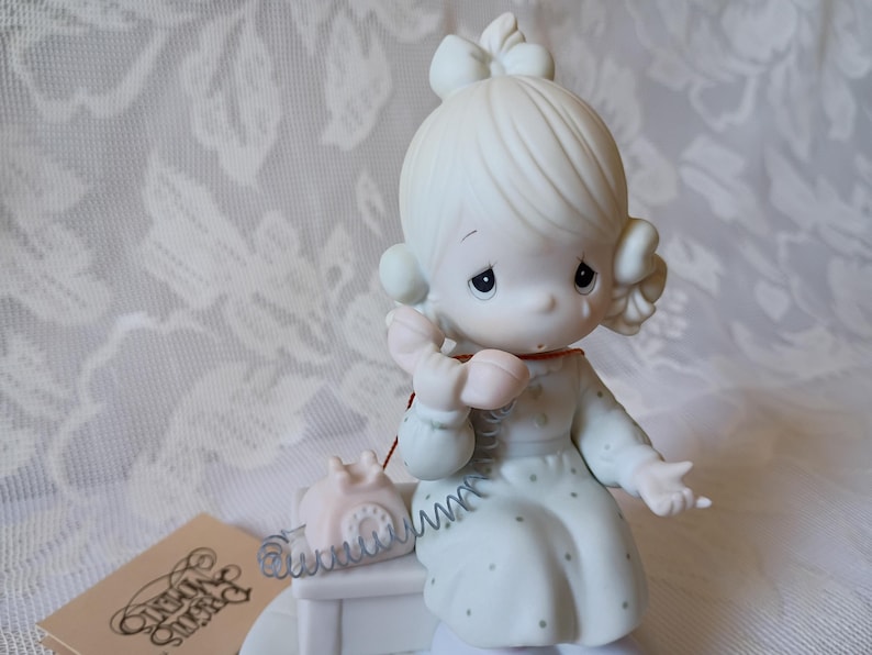 May include: A porcelain figurine of a girl with a bow in her hair, holding a pink telephone receiver. She is wearing a light blue dress with small polka dots. A small pink telephone sits on a white table. The figurine is part of the Precious Moments collection.