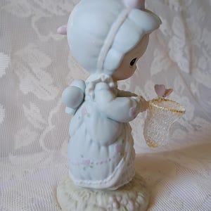 May include: A porcelain figurine of a child in a light blue dress and bonnet, holding a butterfly net. The figurine has pink accents and is standing on a grassy base. A small pink butterfly is near the net.