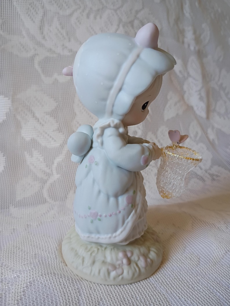 May include: A porcelain figurine of a child in a light blue dress and bonnet, holding a butterfly net. The figurine has pink accents and is standing on a grassy base. A small pink butterfly is near the net.