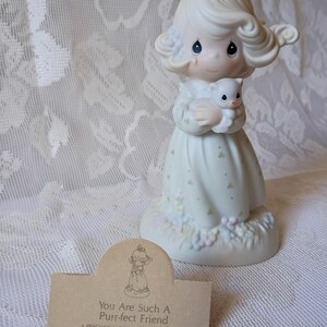 May include: A Precious Moments porcelain figurine of a girl holding a kitten. The figurine is dressed in a white dress with floral accents. The base has pastel-colored flowers. A brown tag reads "You Are Such A Purr-fect Friend."