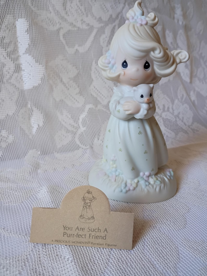 May include: A Precious Moments porcelain figurine of a girl holding a kitten. The figurine is dressed in a white dress with floral accents. The base has pastel-colored flowers. A brown tag reads "You Are Such A Purr-fect Friend."