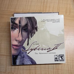 May include: Syberia II PC game box. The box features a woman's face with falling snow. The game title is in a stylized font. The text "The Adventure Continues" is below the title. The game is rated Teen.