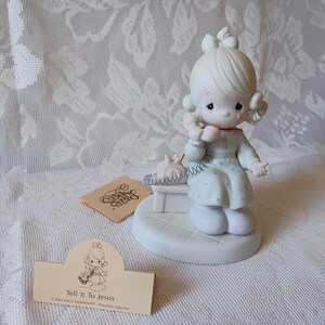 May include: A Precious Moments figurine of a girl with blonde hair, wearing a light blue dress, holding a pink telephone. The figurine is on a white base, with a small tag that reads "Tell It To Jesus".