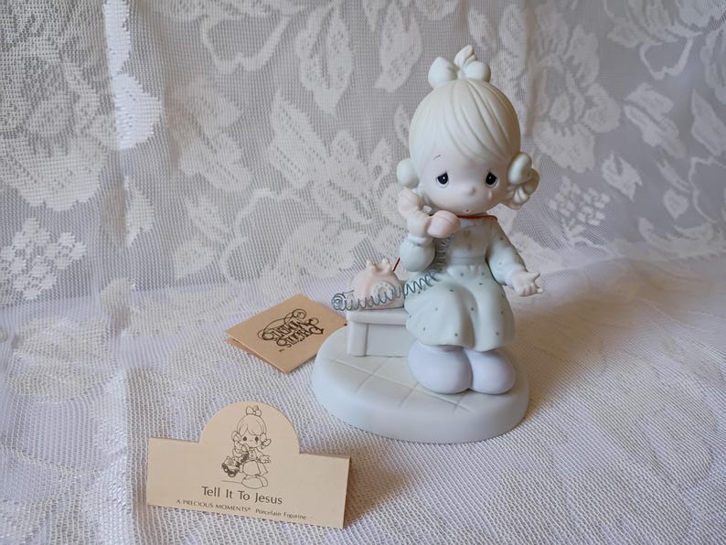 May include: A Precious Moments figurine of a girl with blonde hair, wearing a light blue dress, holding a pink telephone. The figurine is on a white base, with a small tag that reads "Tell It To Jesus".