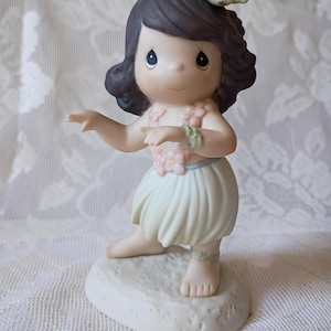 May include: A porcelain figurine of a young girl in a hula outfit. She wears a flower lei, wrist and ankle bracelets, and a flower in her hair. Her skirt is light green, and she is posed in a dance. The figurine is on a white base.