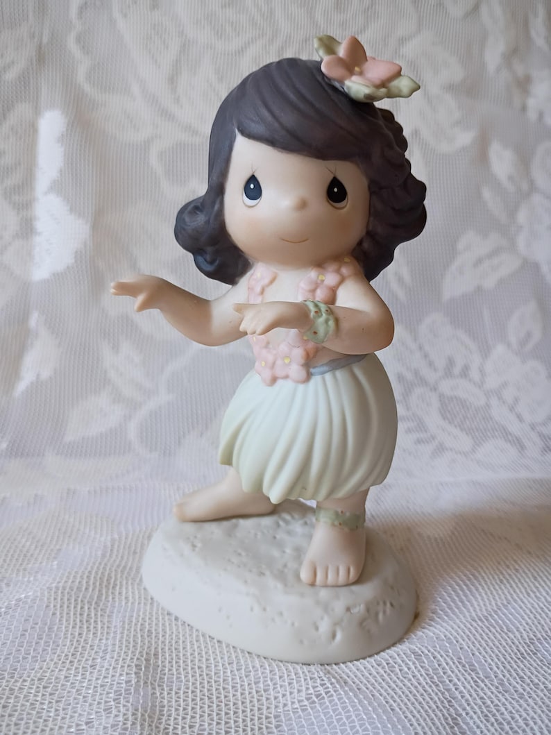 May include: A porcelain figurine of a young girl in a hula outfit. She wears a flower lei, wrist and ankle bracelets, and a flower in her hair. Her skirt is light green, and she is posed in a dance. The figurine is on a white base.