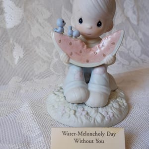 May include: A porcelain figurine of a child holding a watermelon slice, with two blue birds perched on top. The figurine is on a base with flowers and the text "Water-Meloncholy Day Without You" on a tag.