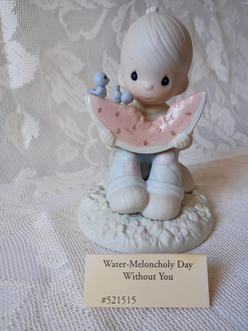 May include: A porcelain figurine of a child holding a watermelon slice, with two blue birds perched on top. The figurine is on a base with flowers and the text "Water-Meloncholy Day Without You" on a tag.