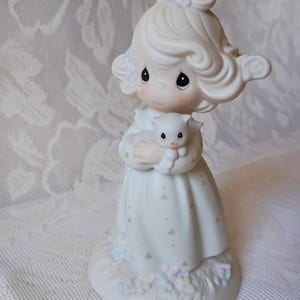 May include: A porcelain figurine of a girl holding a small kitten. The girl wears a white dress with green and pastel floral accents. Her hair is styled with soft curls and adorned with pastel flowers. The figurine is on a base with pastel floral details.