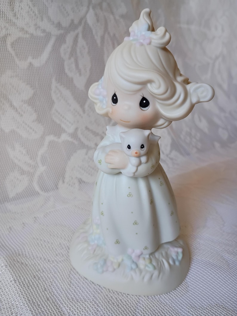 May include: A porcelain figurine of a girl holding a small kitten. The girl wears a white dress with green and pastel floral accents. Her hair is styled with soft curls and adorned with pastel flowers. The figurine is on a base with pastel floral details.