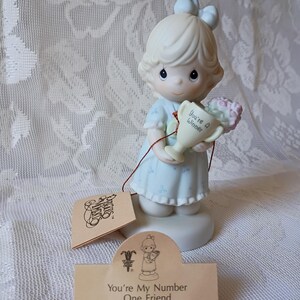 May include: A Precious Moments porcelain figurine of a girl holding a trophy and flowers. The trophy reads "You're a Winner." The figurine is wearing a light blue dress and has a matching bow. The tag reads "You're My Number One Friend."
