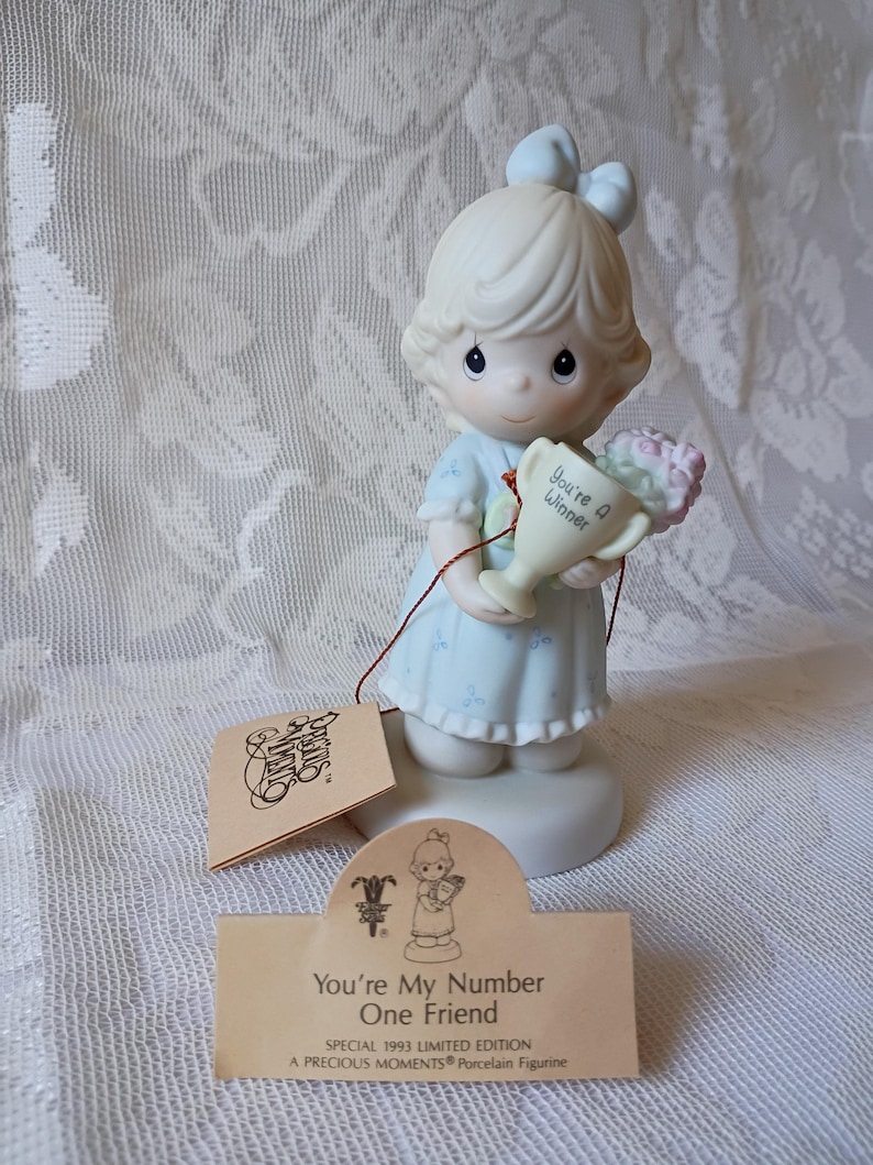 May include: A Precious Moments porcelain figurine of a girl holding a trophy and flowers. The trophy reads "You're a Winner." The figurine is wearing a light blue dress and has a matching bow. The tag reads "You're My Number One Friend."