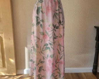May include: A long, flowing skirt with a light pink background and a floral pattern. The pattern features pink roses and green leaves. The skirt is made of a lightweight fabric and has a subtle sheen. The skirt is displayed on a mannequin.