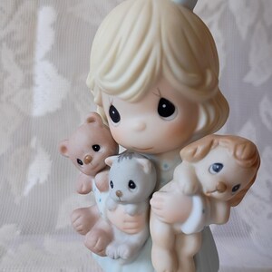 May include: A porcelain figurine of a girl holding a teddy bear, a kitten, and a puppy. The girl has blonde hair and a blue bow with white polka dots. The figurine is painted in soft pastel colors, with a light blue dress and a white base.