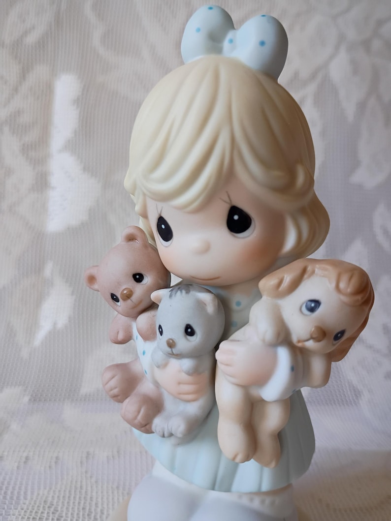 May include: A porcelain figurine of a girl holding a teddy bear, a kitten, and a puppy. The girl has blonde hair and a blue bow with white polka dots. The figurine is painted in soft pastel colors, with a light blue dress and a white base.
