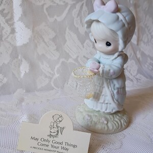 May include: A Precious Moments porcelain figurine of a child in a bonnet holding a net. The figurine is light blue and white, with a pink bow. A card reads "May Only Good Things Come Your Way."