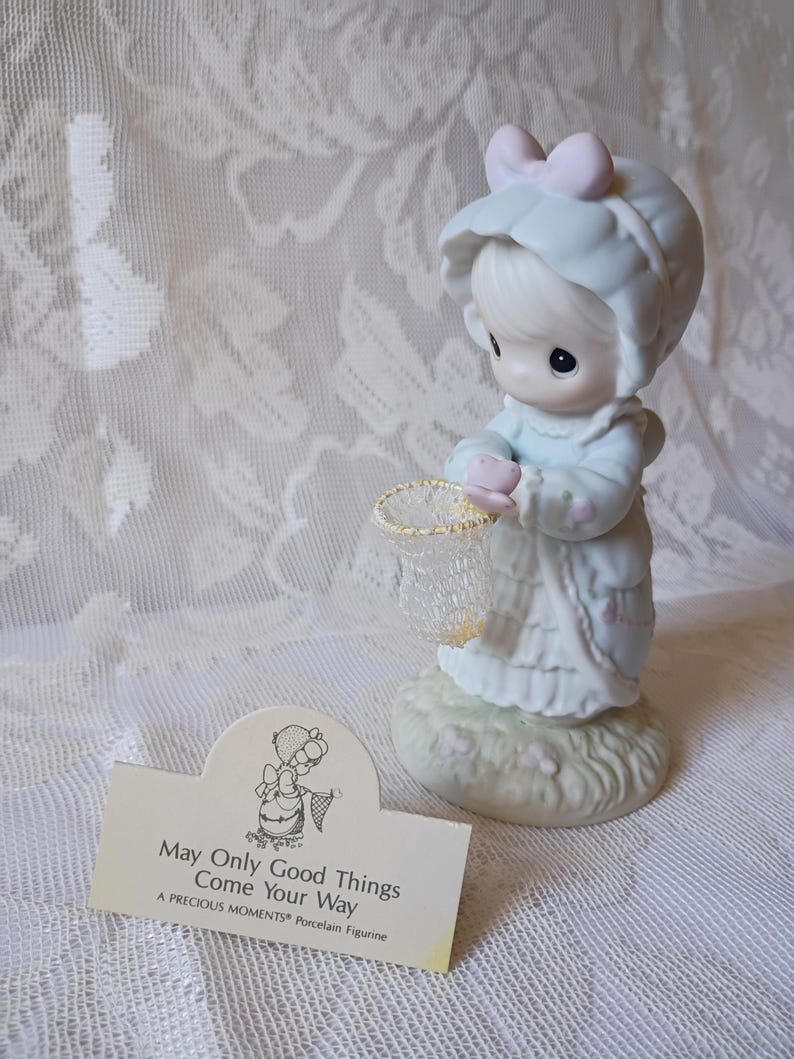 May include: A Precious Moments porcelain figurine of a child in a bonnet holding a net. The figurine is light blue and white, with a pink bow. A card reads "May Only Good Things Come Your Way."