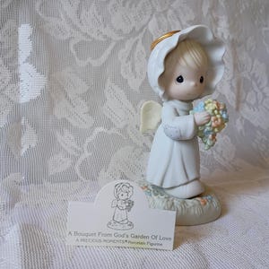 May include: A Precious Moments porcelain figurine of an angel holding a bouquet of pastel-colored flowers. The angel wears a white gown and a halo. The base of the figurine is decorated with flowers and greenery. The text on the card reads: "A Bouquet From God's Garden of Love".