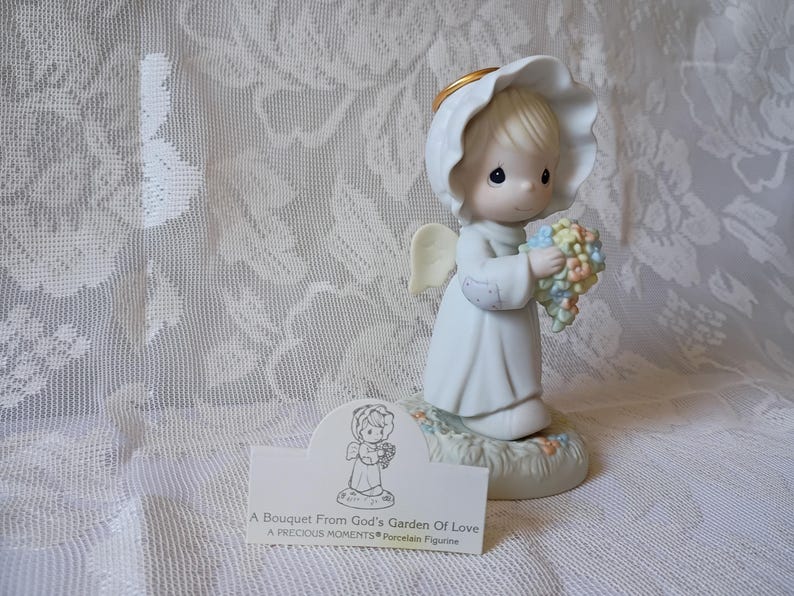 May include: A Precious Moments porcelain figurine of an angel holding a bouquet of pastel-colored flowers. The angel wears a white gown and a halo. The base of the figurine is decorated with flowers and greenery. The text on the card reads: "A Bouquet From God's Garden of Love".