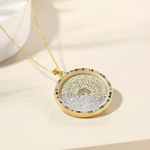 May include: A gold-colored pendant necklace with a round, ornate pendant. The pendant features a detailed design with colorful gemstones around the edge and clear crystals inside. The necklace chain is also gold-colored.