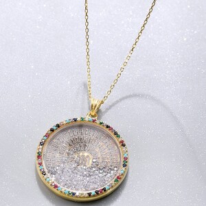 May include: Gold-toned necklace with a round pendant. The pendant features a detailed design with a central element surrounded by clear and colorful gemstones. The chain is delicate, and the pendant hangs gracefully.