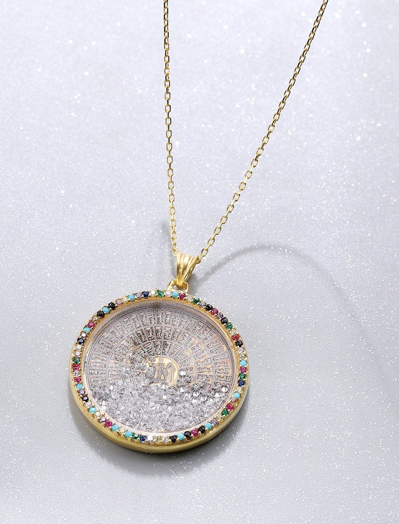 May include: Gold-toned necklace with a round pendant. The pendant features a detailed design with a central element surrounded by clear and colorful gemstones. The chain is delicate, and the pendant hangs gracefully.