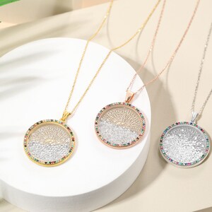 May include: Three pendant necklaces with round, tree-of-life designs. One is gold-toned, one rose gold, and one silver. Each pendant has a colorful gemstone border and hangs on a delicate chain. The necklaces are displayed on a white, circular platform.